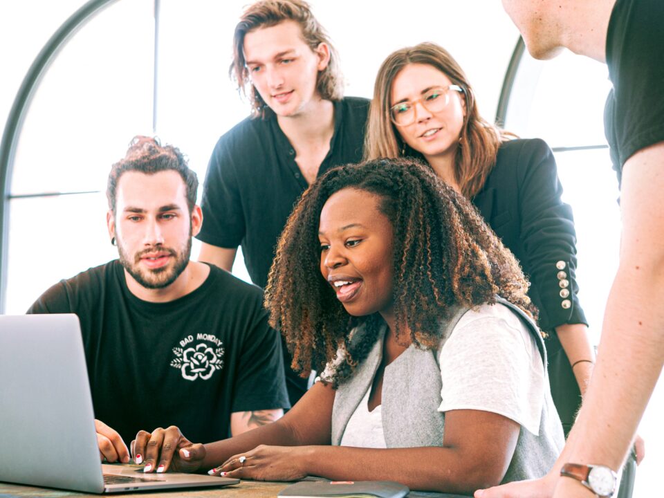 A woman shows something on the laptop, and three men and one woman look at the screen